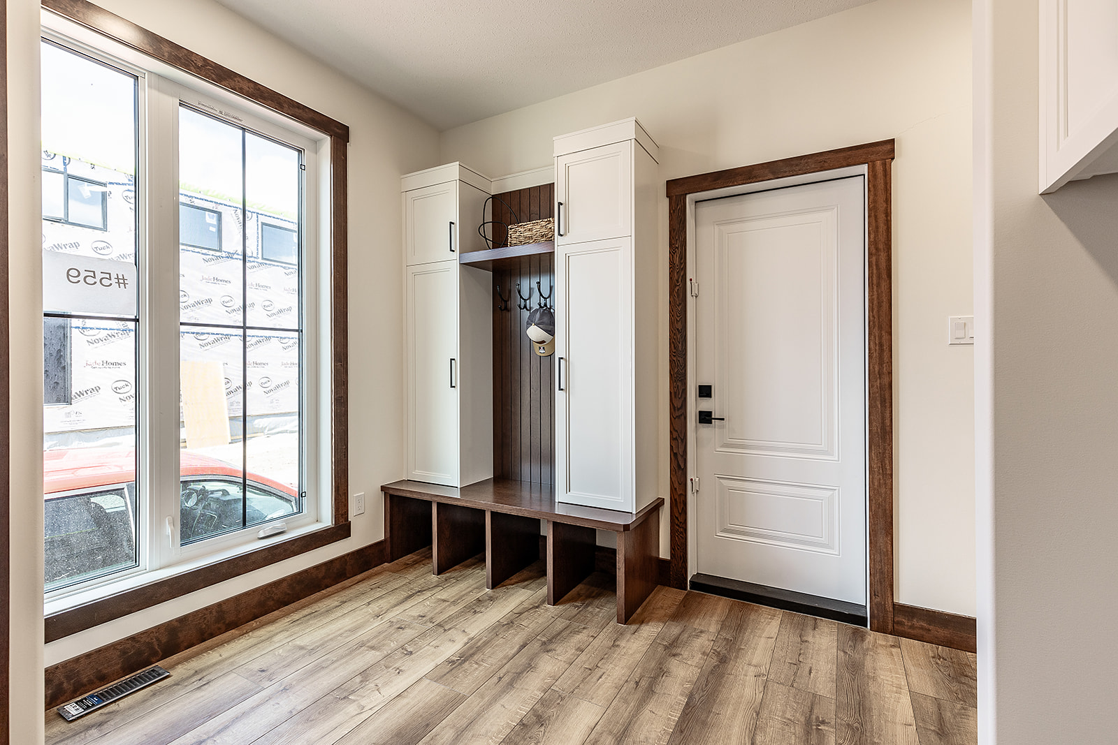 Modern mudroom with light wood flooring, white storage cabinets, bench seating with cubbies, and coat hooks. Large window offers natural light.