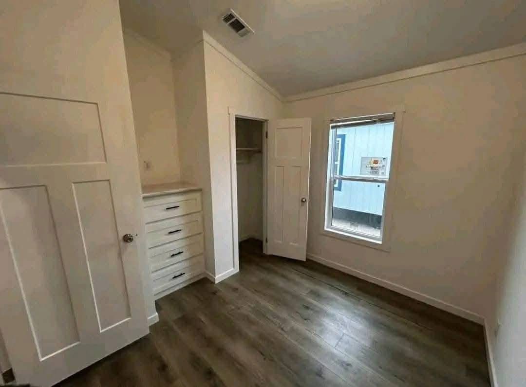A small, empty room with light-colored walls and wood floors. It features a window on the right, built-in drawers on the left, and a closet with an open door.