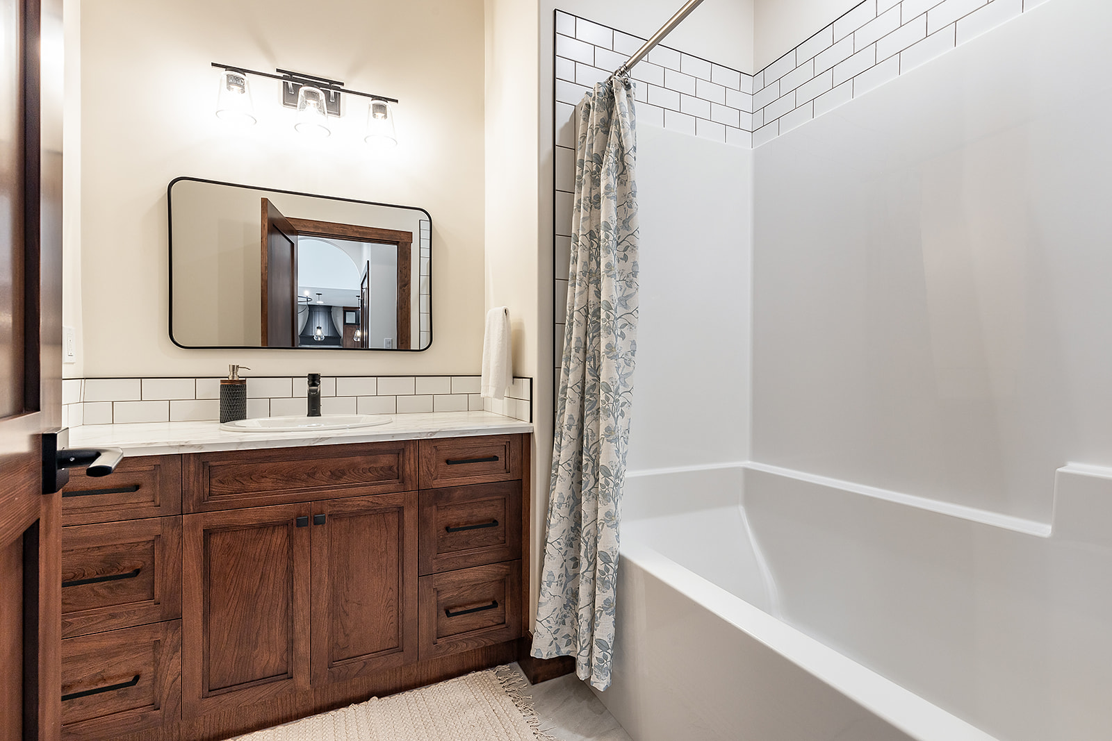 A modern bathroom with a wood vanity, white countertop, and large mirror under bright lights. A tiled shower with a patterned curtain is to the right.