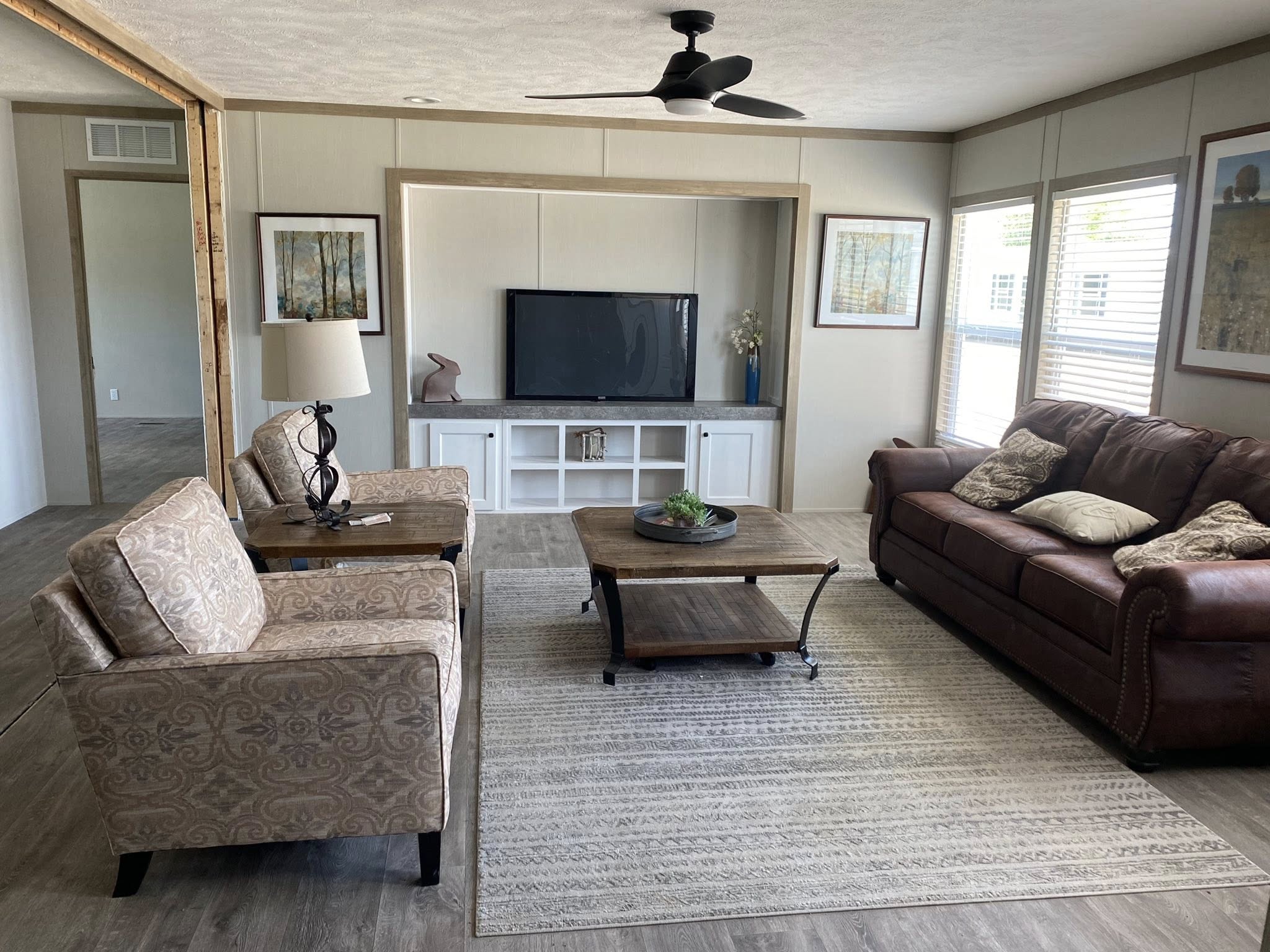 Cozy living room with a brown leather sofa, patterned armchairs, coffee table on a large rug, flat-screen TV, framed artwork, and large windows.