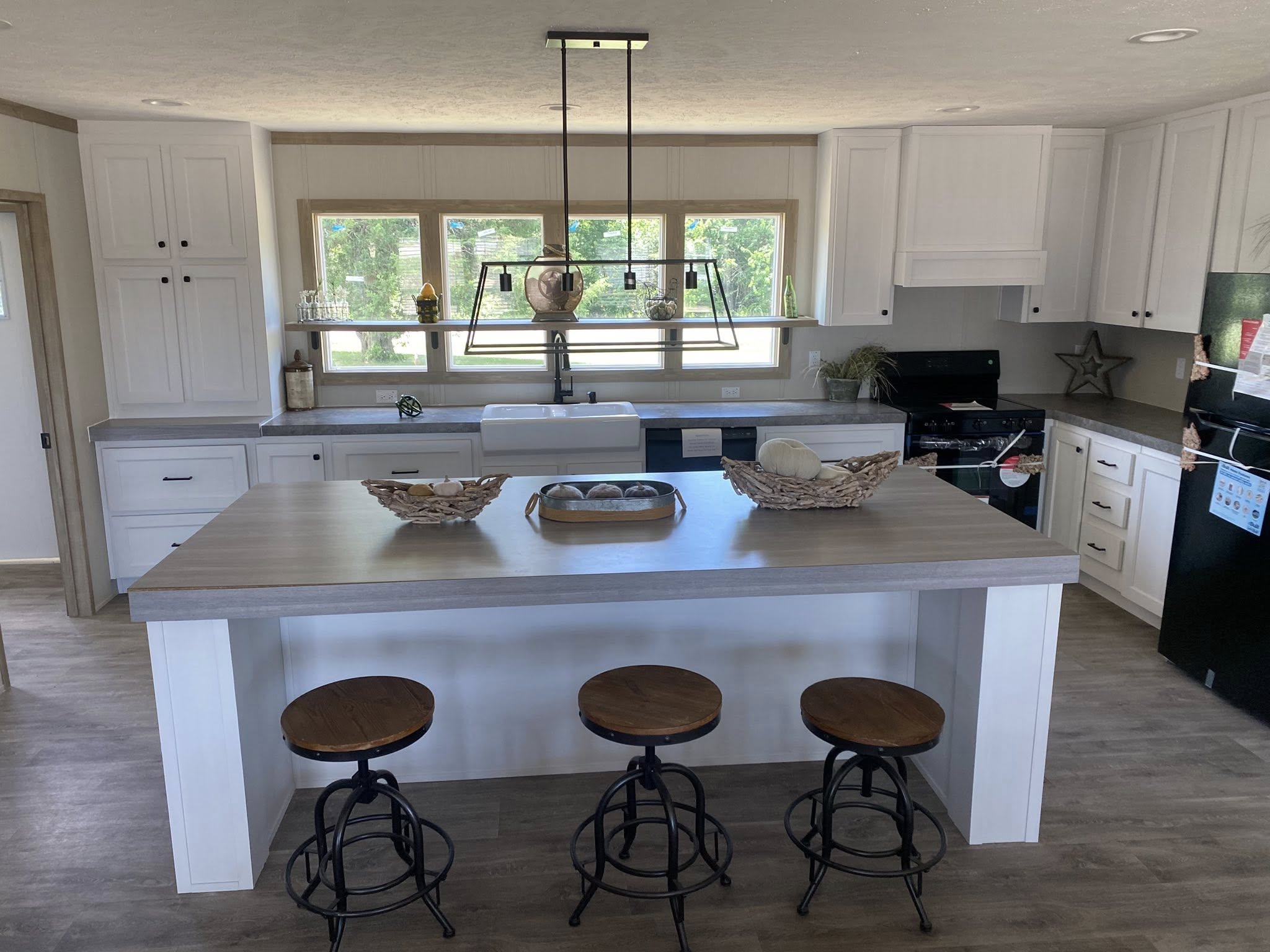 Modern kitchen with a large island featuring wooden stools, white cabinets, and a farmhouse sink. Bright windows and neutral tones create an inviting atmosphere.