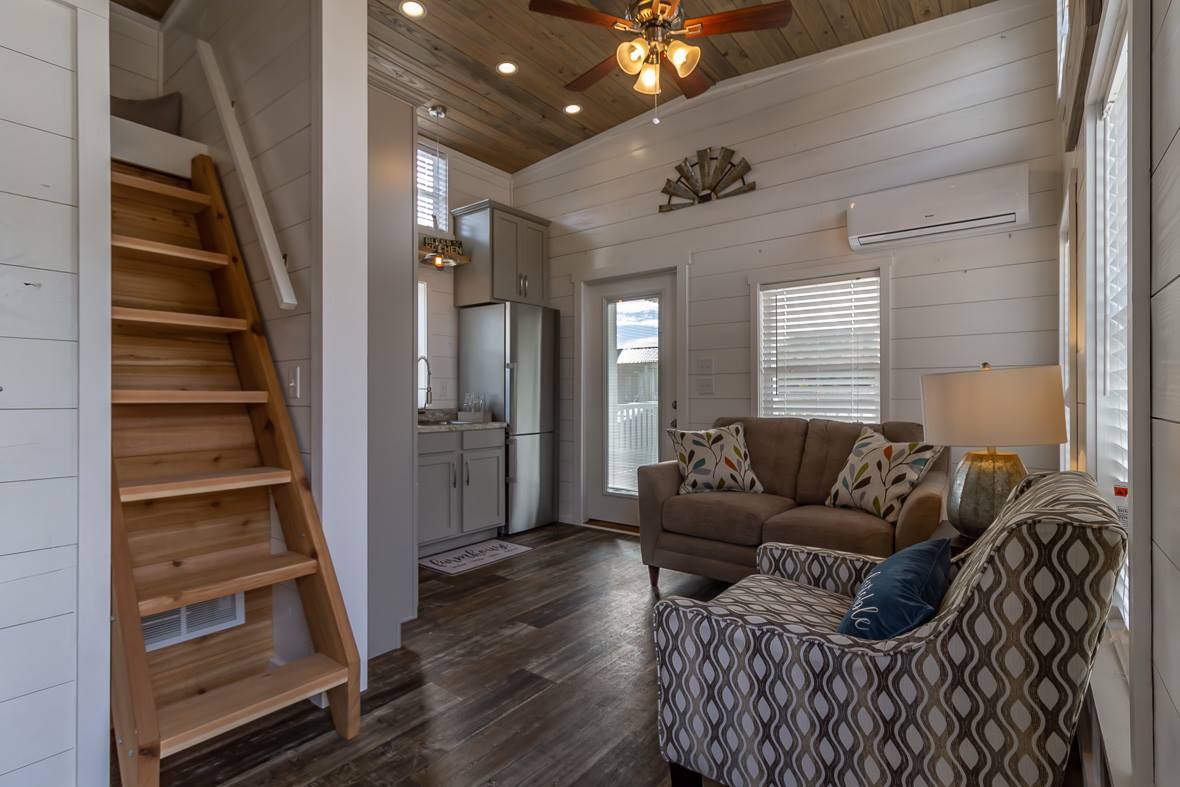Cozy tiny house interior with wooden stairs, compact kitchen with gray cabinets, a plush sofa, patterned armchair, and warm lighting creating a homely feel.