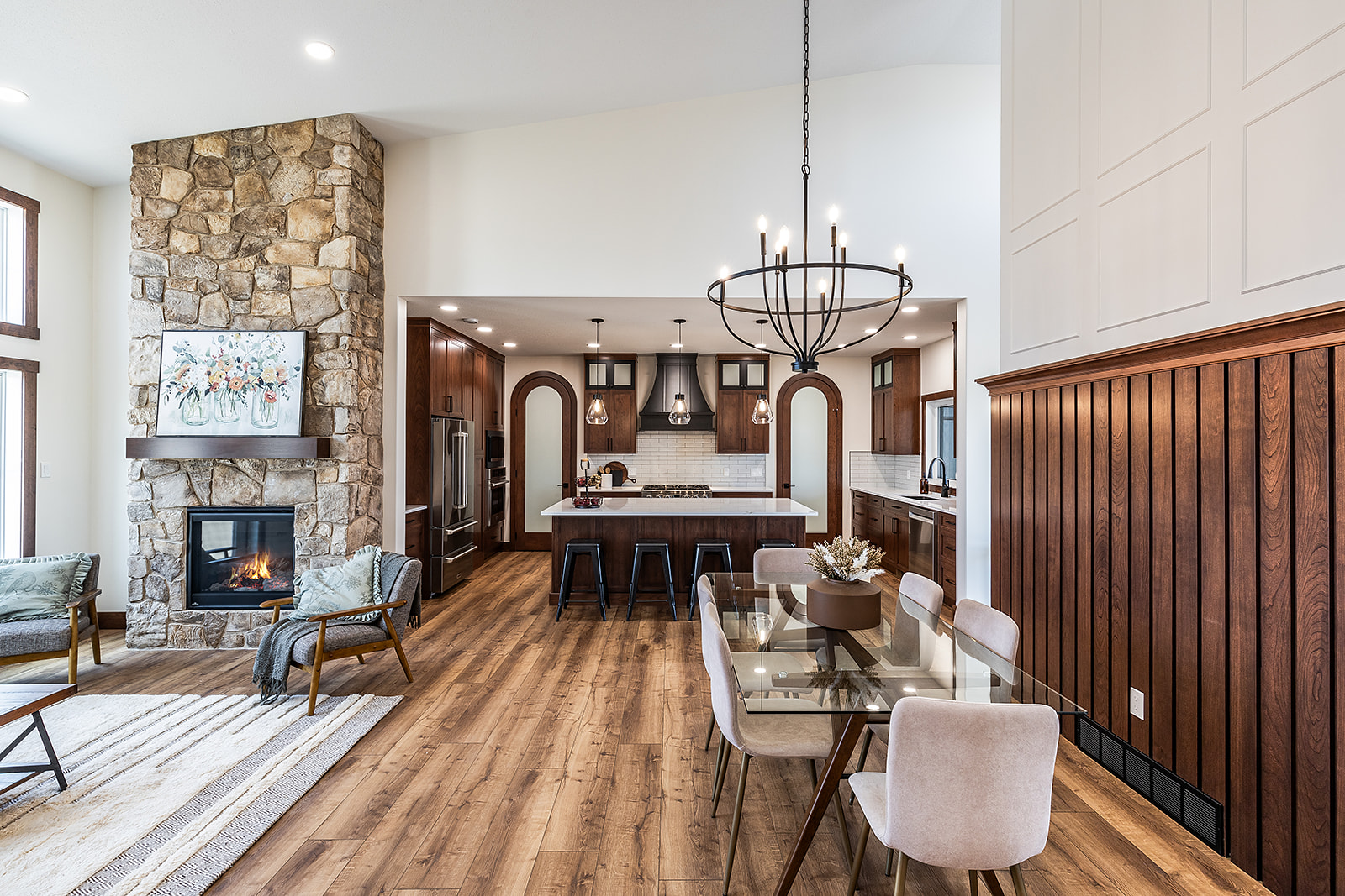 Modern open-concept kitchen and dining area with wooden floors, stone fireplace, and a large chandelier. Warm tones create an inviting atmosphere.