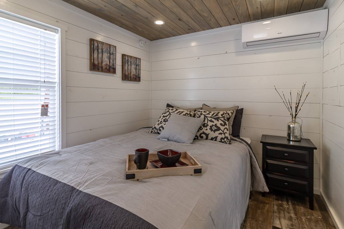 Cozy bedroom with shiplap walls, wooden ceiling, and a soft bed. A tray with cups sits on the bed, and a vase adorns the nightstand. Warm, inviting ambiance.