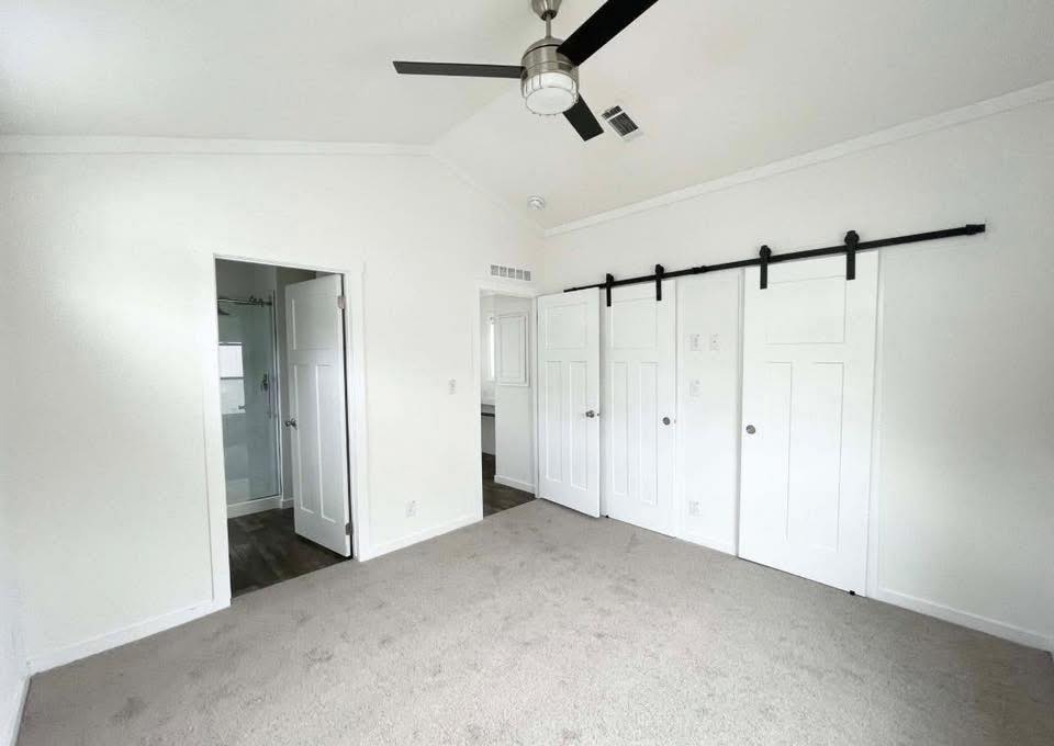 Bright, empty room with white walls and carpet, featuring a ceiling fan and barn-style closet doors; an open bathroom door to the left.