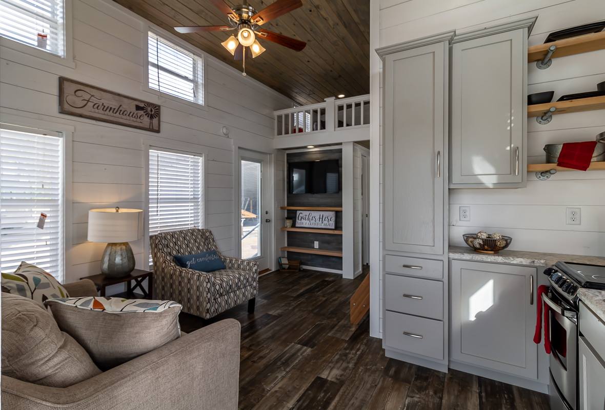 Cozy living room with modern farmhouse decor, featuring a patterned armchair, beige sofa, wooden floor, vaulted wood ceiling, and "Farmhouse" sign.