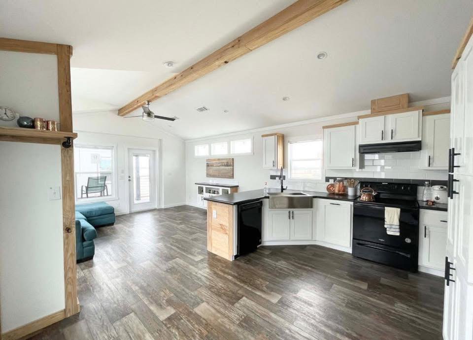 Spacious kitchen with white cabinets and black appliances, featuring a farmhouse sink and wooden accents. Cozy sitting area with teal sofas. Bright, airy atmosphere.