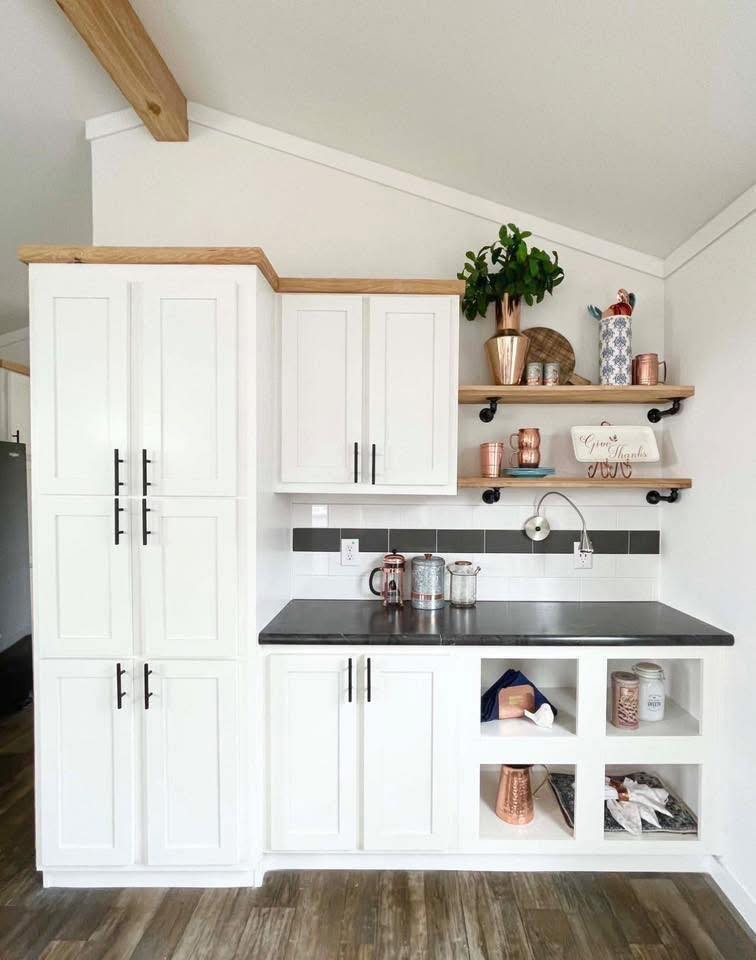 A modern kitchen nook features white cabinets, black handles, and a dark countertop. Wooden shelves hold copper containers and a plant, creating a cozy vibe.