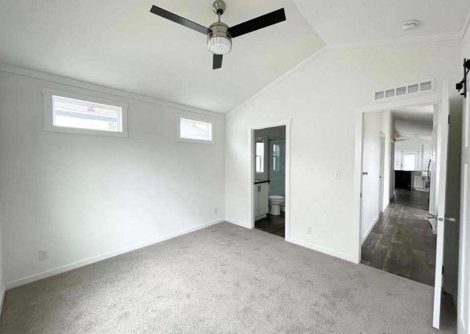 Bright, unfurnished bedroom with white walls, carpeted floor, ceiling fan, two small windows, and a doorway leading to a bathroom and hallway.