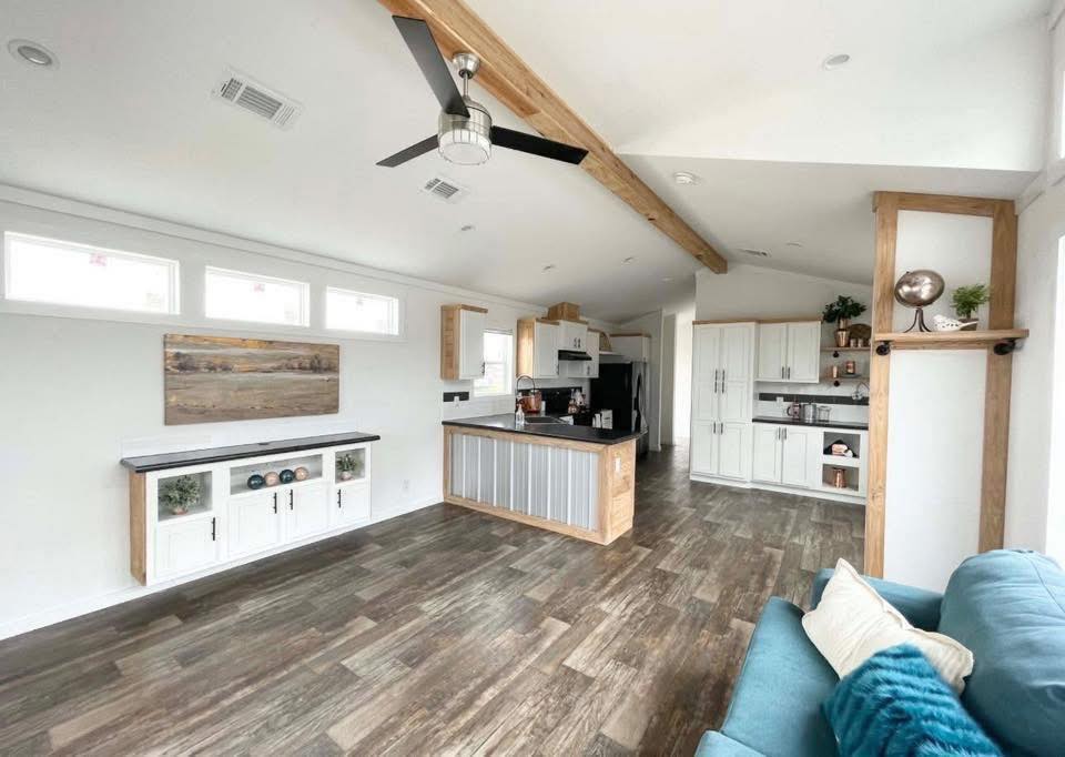 Spacious modern kitchen with wooden floors, white cabinets, and a central island. Bright with natural light, complemented by a ceiling fan and decor.