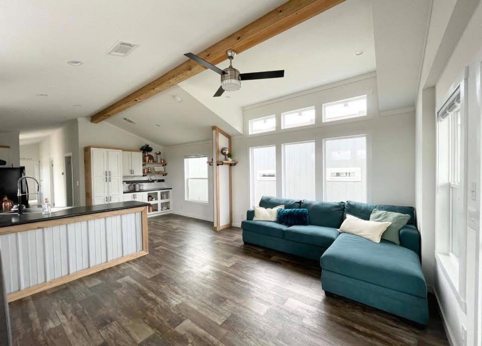Spacious, modern living room with a teal sectional sofa, wood accents, large windows, and ceiling fan. Light wood flooring and an airy atmosphere enhance the room.