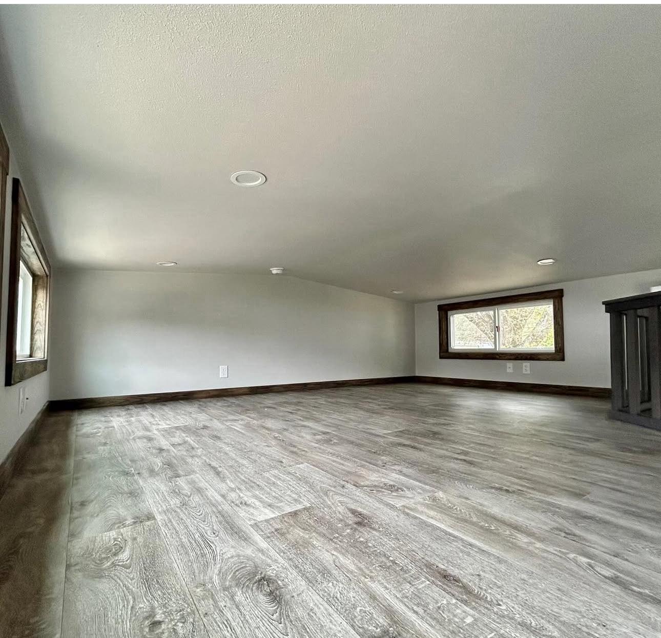 Spacious attic with sloped ceiling, featuring light gray wood flooring and neutral walls. Windows provide natural light, creating a calm atmosphere.