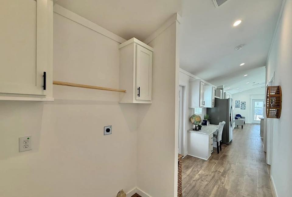 Bright, modern interior with wood flooring and white walls. A laundry nook with cabinets is on the left, leading to a kitchen with stainless steel appliances.