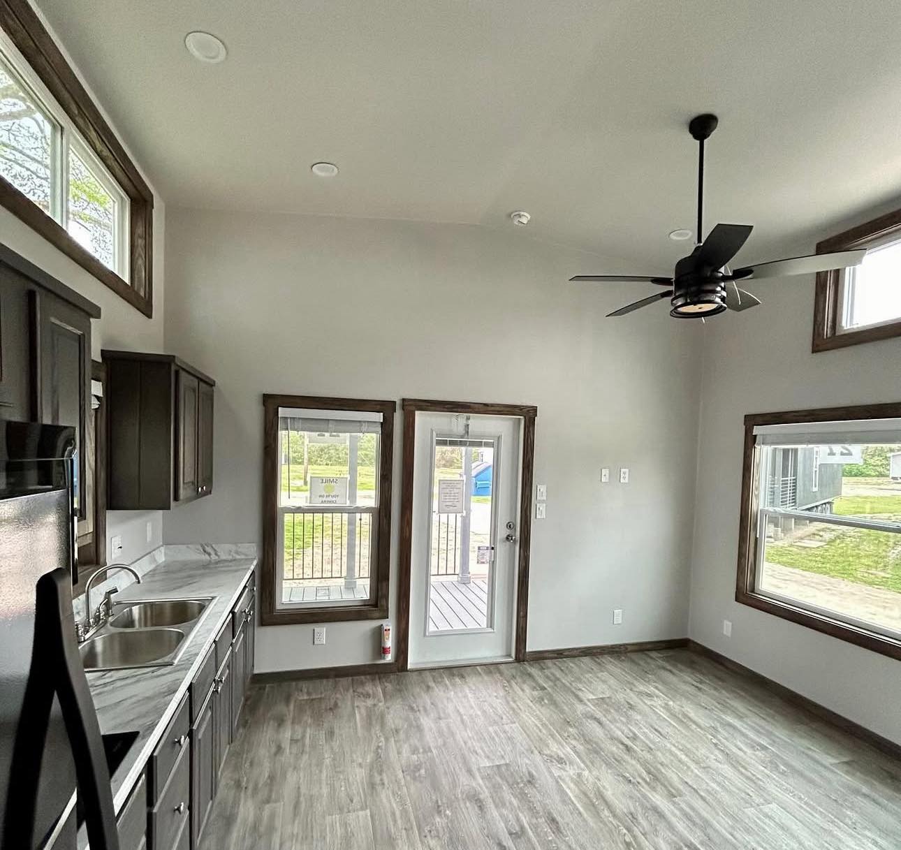Modern kitchen with dark cabinets, marble countertops, and stainless steel sink. Bright with large windows, gray flooring, and ceiling fan for an airy feel.