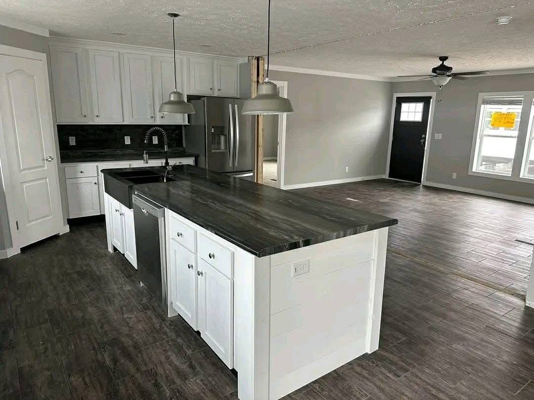 Modern kitchen with white cabinets, dark countertops, and stainless steel appliances. An island with pendant lights overlooks an open, bright living area.