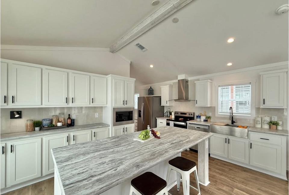 Modern kitchen with white cabinets, a large gray marble island, and bar stools. Stainless steel appliances and a window bringing in natural light. Cozy and inviting ambiance.