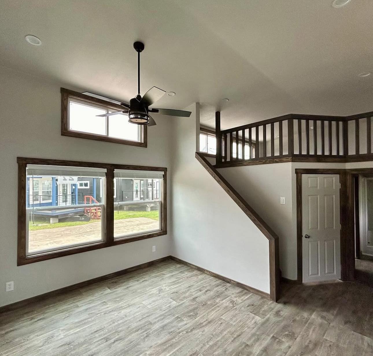 Spacious room with large windows, loft area with railing, wood flooring, and ceiling fan. Sunlight streams in, creating a bright, airy atmosphere.