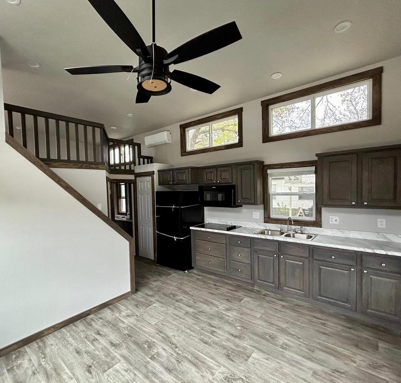 Modern tiny home interior with a loft, dark cabinetry, marble countertops, and wooden flooring. Large ceiling fan and high windows add open ambiance.