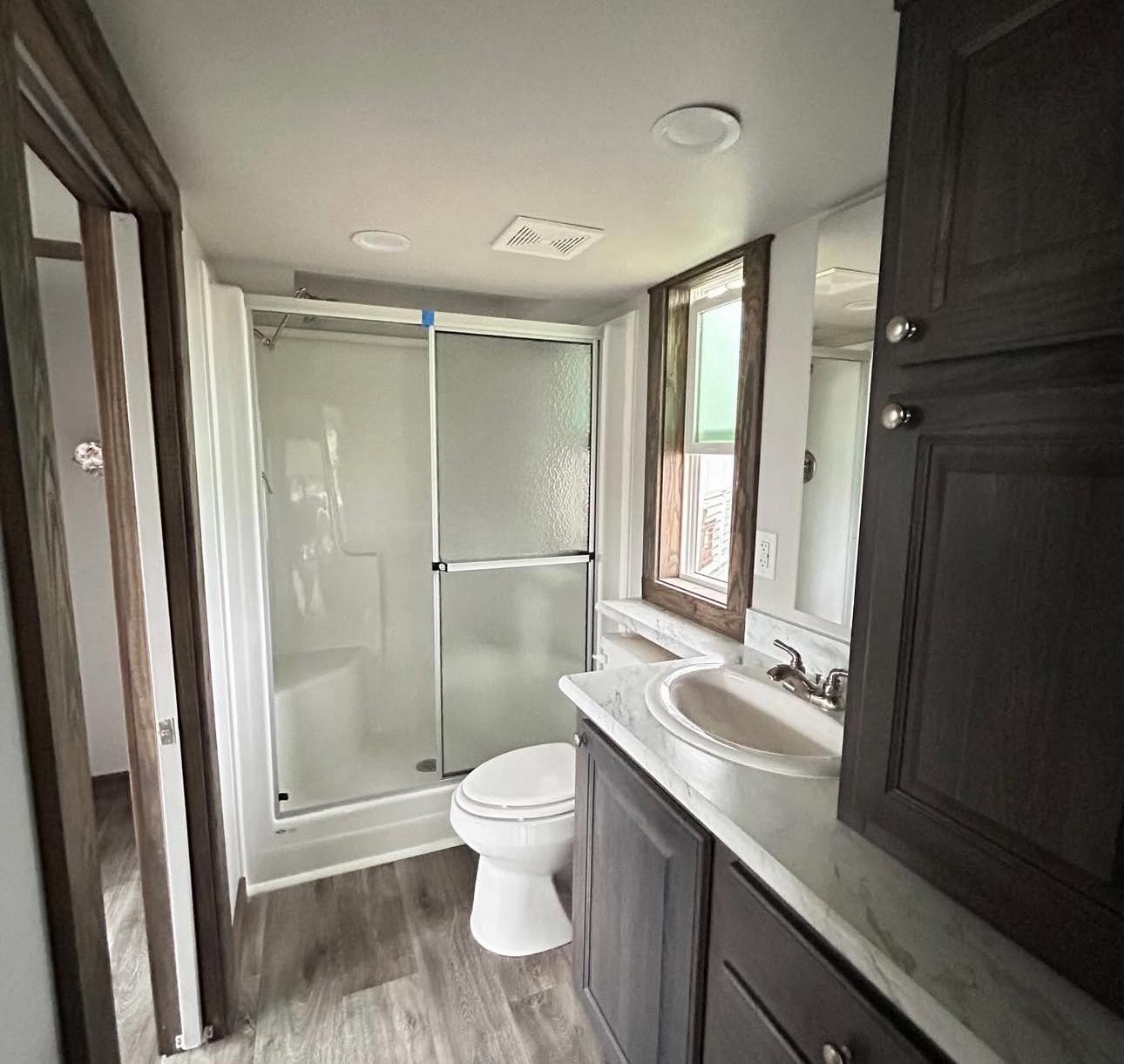 A small bathroom features a shower with frosted glass doors, a toilet, and a sink with a marble countertop. Dark wooden cabinets create a modern look.