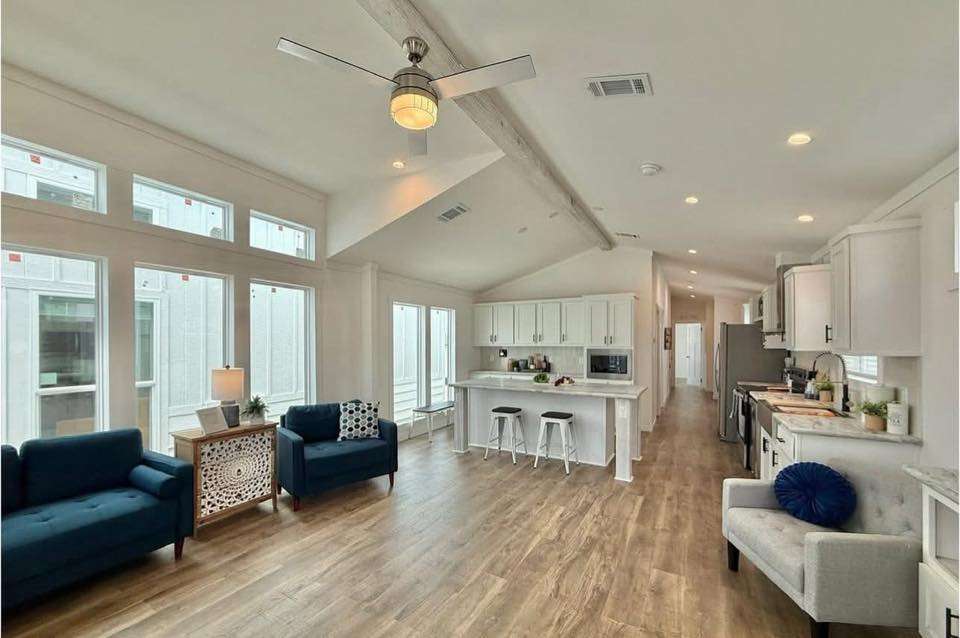 Bright, modern open-concept living space with wood floors, featuring navy sofas, a ceiling fan, large windows, and a kitchen with white cabinets.