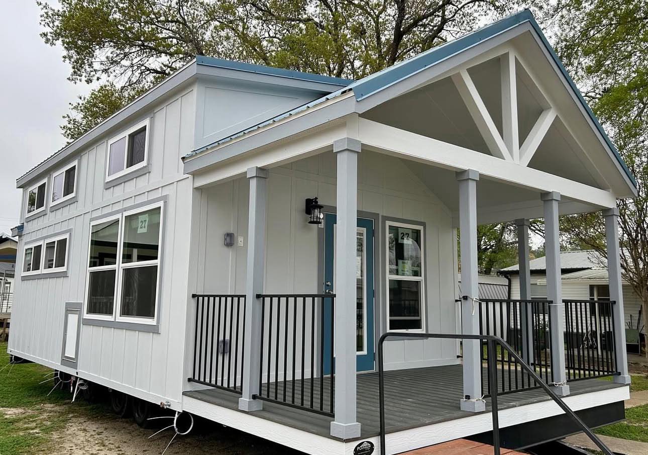 A modern tiny house with white siding and blue accents stands in a green, leafy area. It features large windows and a small porch with black railings.