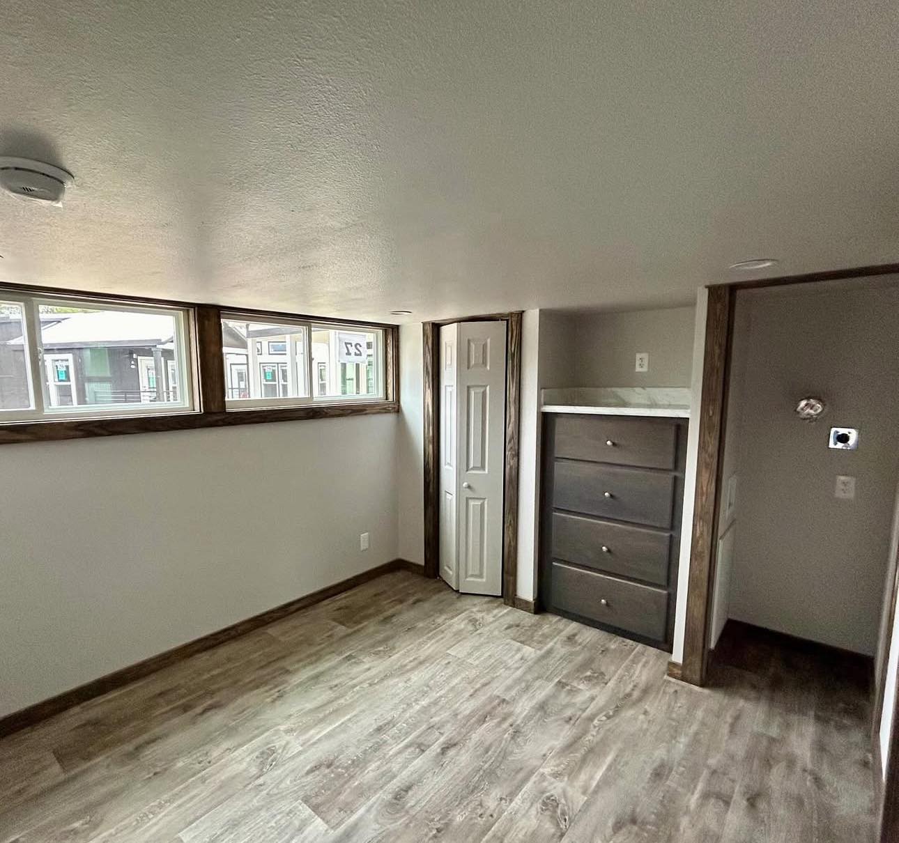 A small room with light wood flooring, recessed lights, and a row of high windows. It features a built-in dark wood dresser and a narrow closet with a white door.