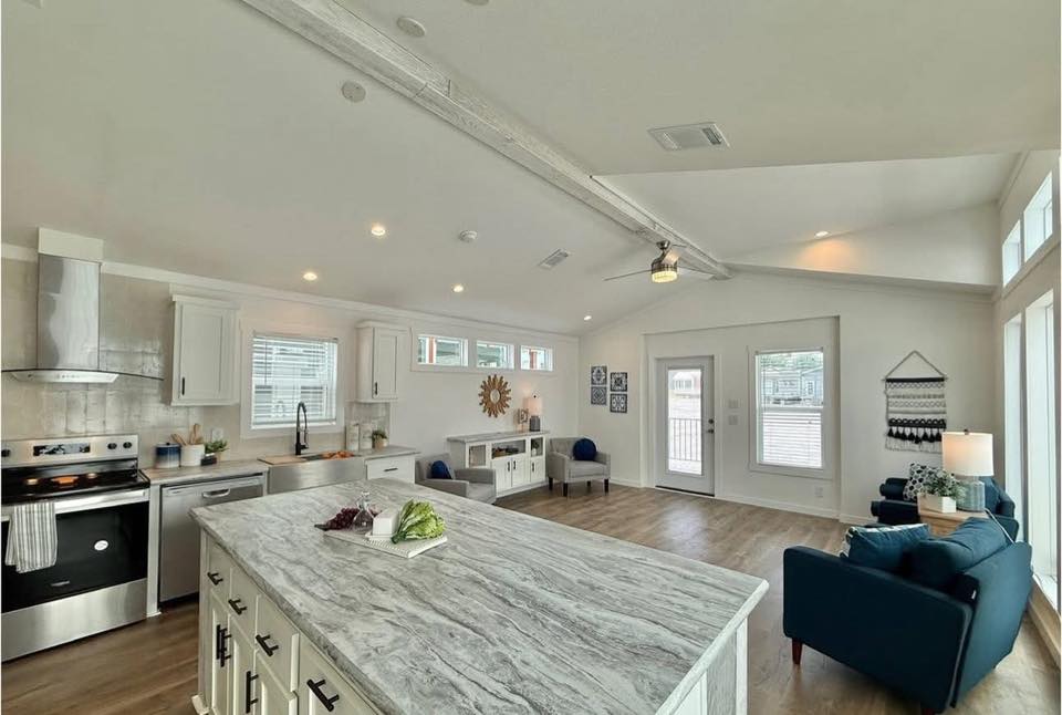 Spacious open-concept living area with a modern kitchen and island, wooden floors, and blue seating. Bright natural light and neutral decor create a welcoming ambiance.