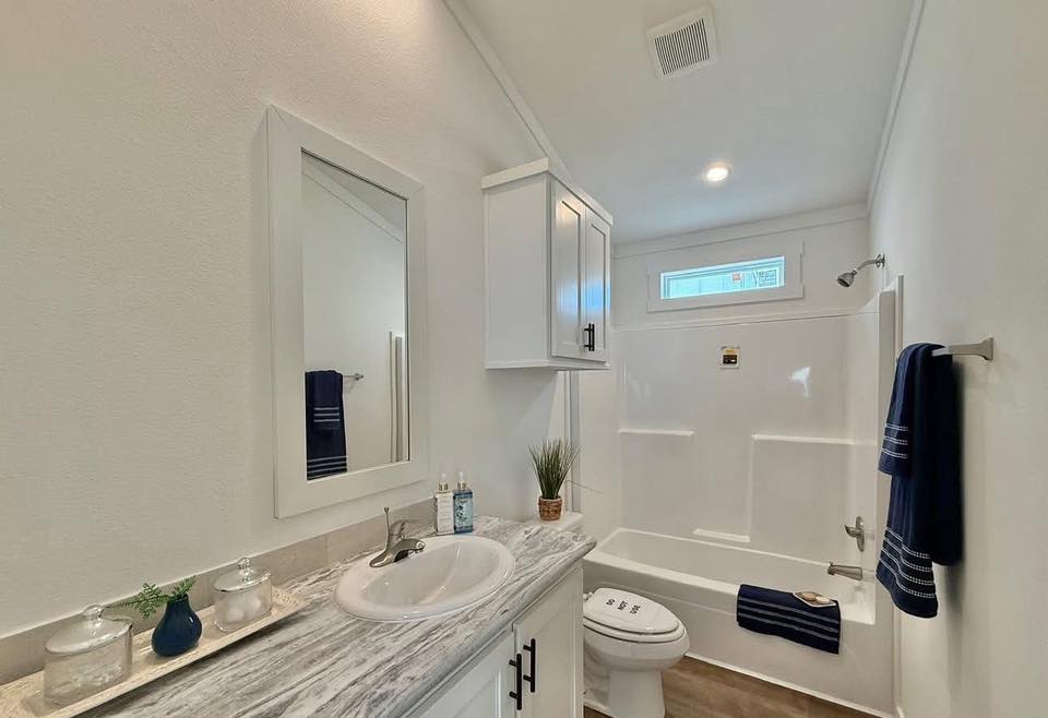A modern bathroom featuring a marble countertop with a sink, white cabinets, and decorative items. The space includes a toilet, bathtub, and navy towels. Bright, clean, and minimalist.
