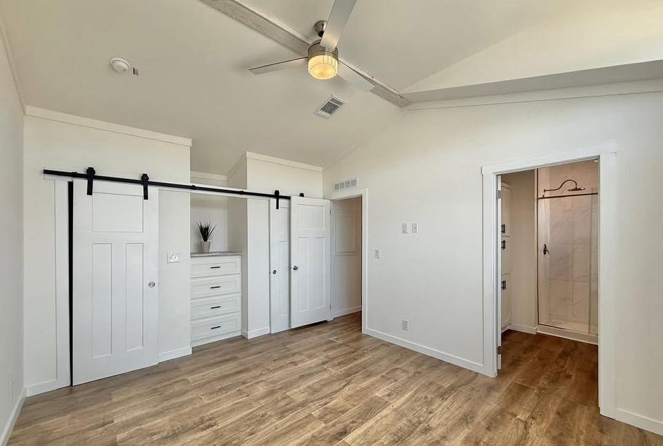 Bedroom with wooden floor, white walls, and a ceiling fan. Features sliding barn doors concealing a closet and dresser. An adjacent doorway leads to a tiled shower area. Bright, minimalist, and modern design.