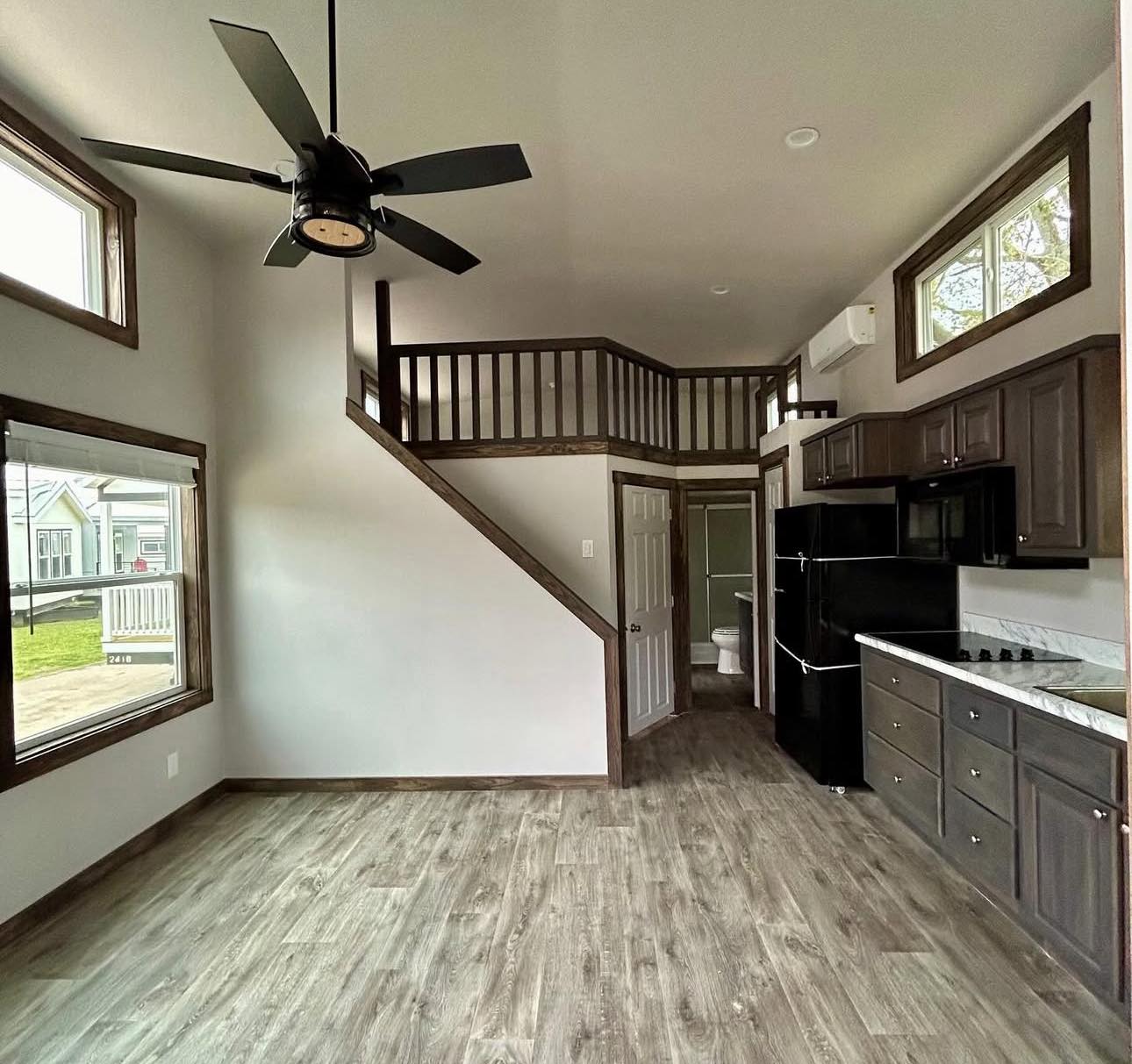 Modern tiny home interior with wood-look flooring, a ceiling fan, and a loft above. Kitchen features dark cabinets and appliances, creating a cozy atmosphere.