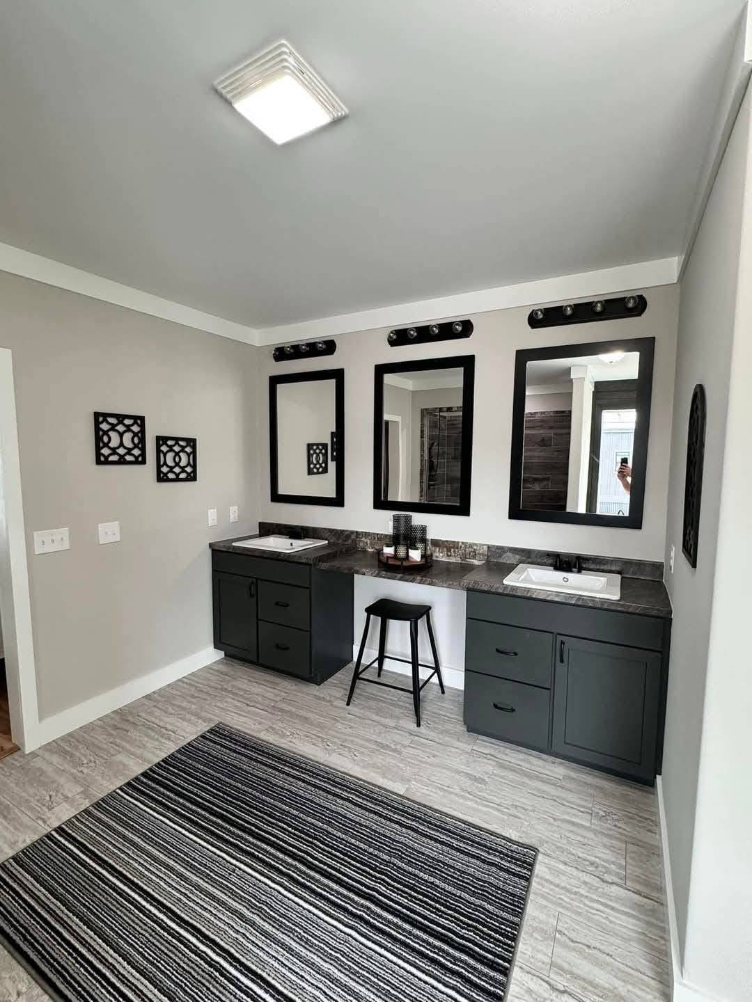 Modern bathroom with dual dark gray vanities, large mirrors, and black decor accents. A striped rug covers the tile floor, creating a sleek, elegant look.