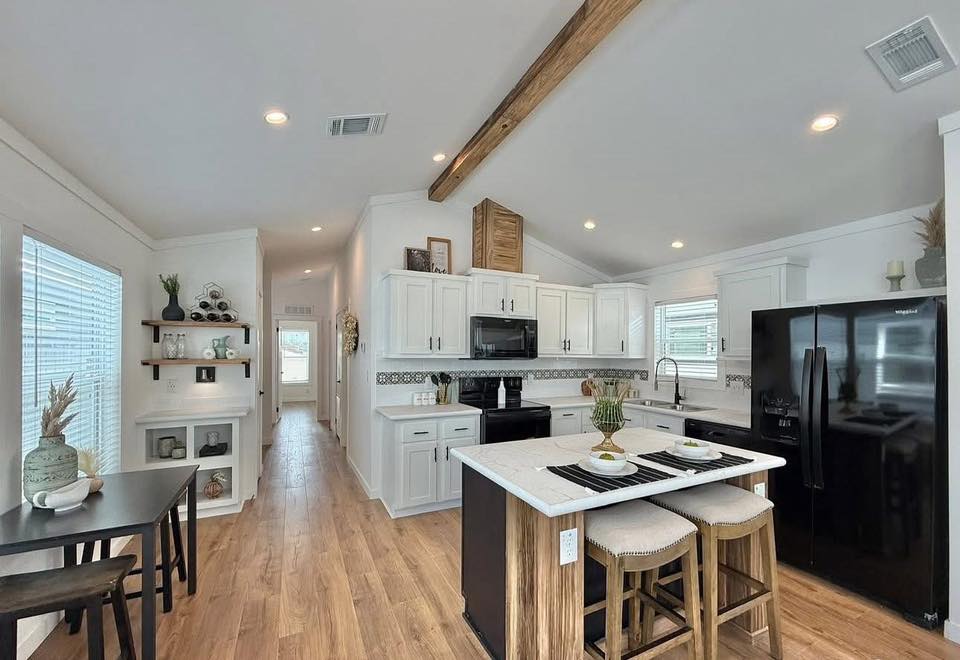 Modern kitchen with white cabinets, wood floors, and a vaulted ceiling featuring a wooden beam. A black fridge matches the stove and microwave. A central island with stools offers seating, while decor on shelves adds a cozy touch.