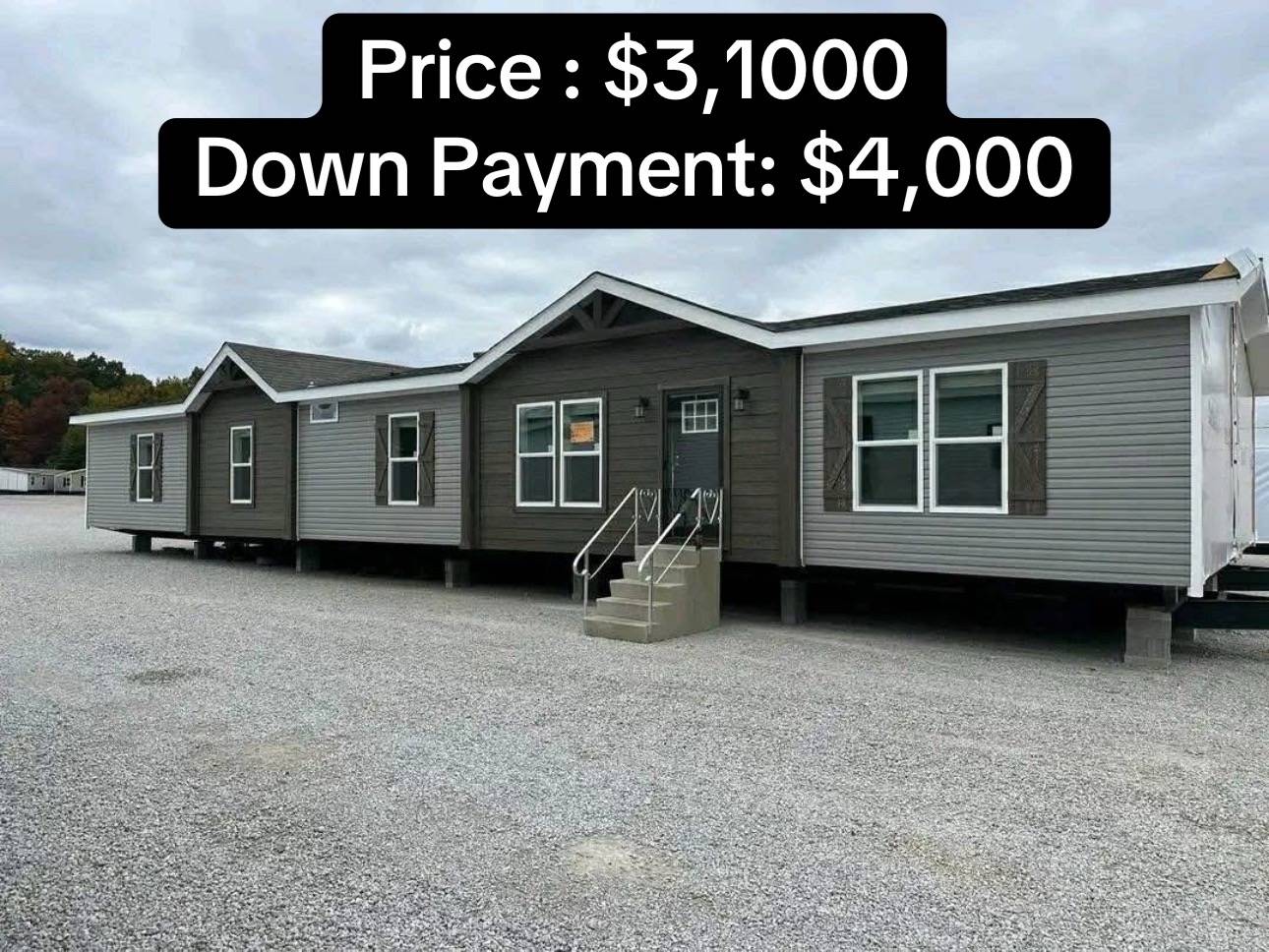 Grey modular home displayed on a gravel lot with small stairs leading to the entrance. Price: $31,000. Down Payment: $4,000. Cloudy sky background.