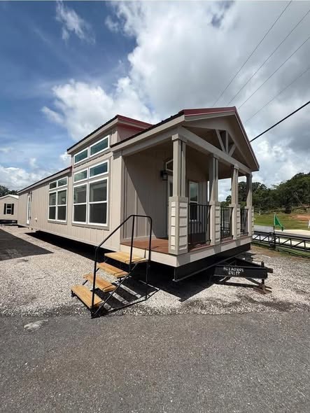 A modern tiny house with beige siding is set on gravel, featuring a small porch and metal steps. Large windows line the side, under a partly cloudy sky.