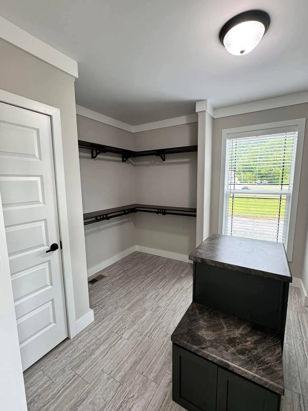 Bright, modern walk-in closet with light gray walls and tile floor. Dark shelving lines two walls, and a large window with blinds lets in natural light.