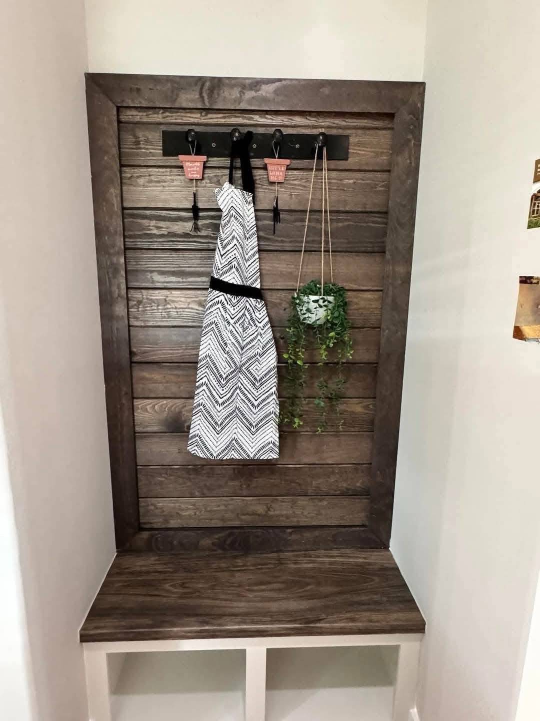 Wooden bench and back panel with hooks holding a patterned apron and a potted plant. Cozy, rustic decor with a hint of greenery.