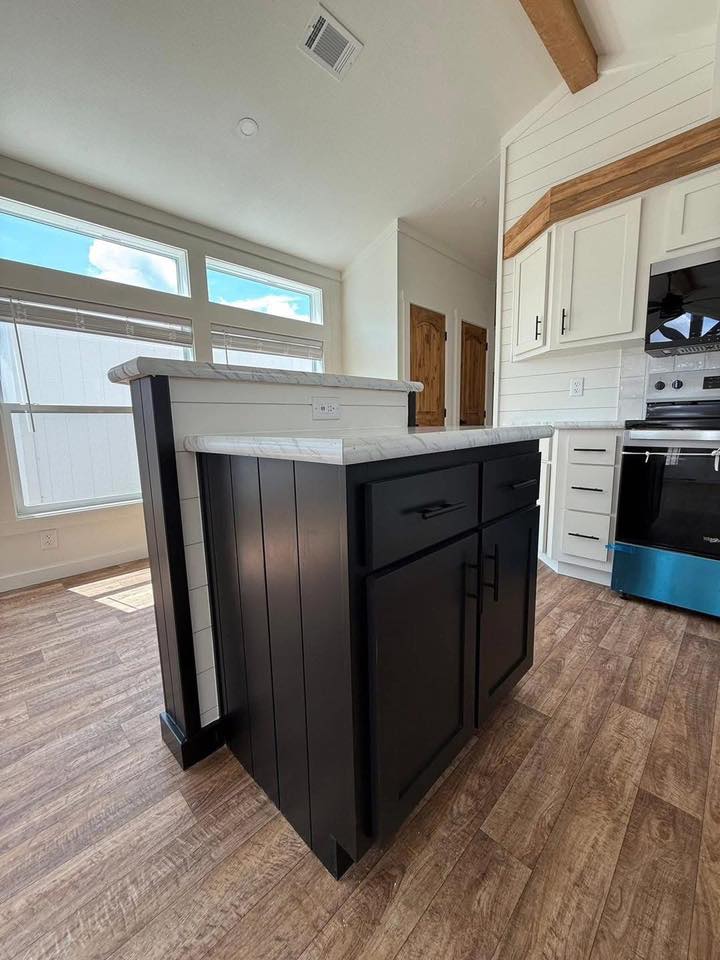 Modern kitchen with wood flooring, a black island with white marble countertop, and white cabinets. Large windows let in natural light.