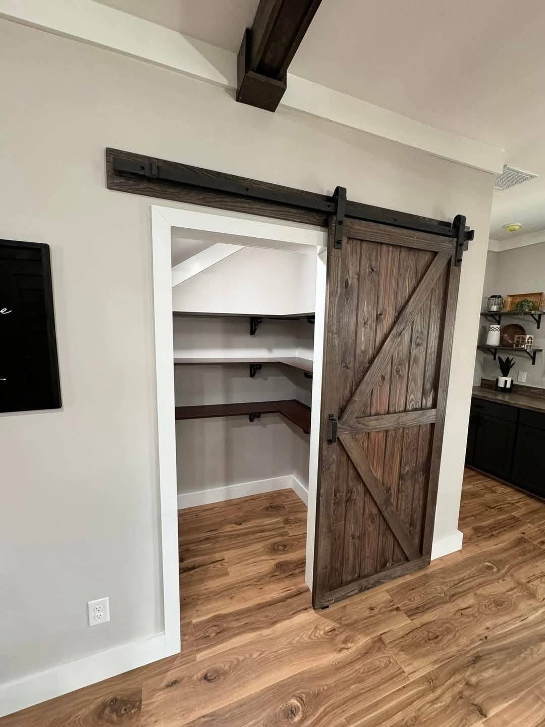 Wooden sliding barn door partially open on a pantry closet with white and wood shelves. Hardwood floors and dark cabinets create a cozy, rustic feel.