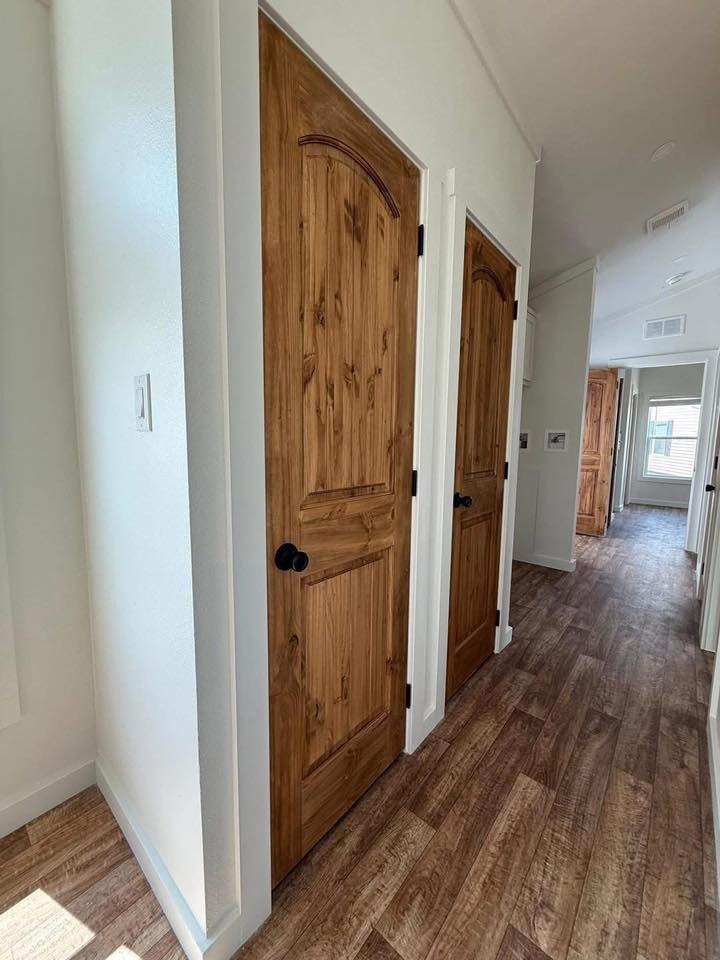 A hallway with light wood flooring features two wooden doors on the left. Sunlight streams through a distant window, creating a warm, cozy atmosphere.
