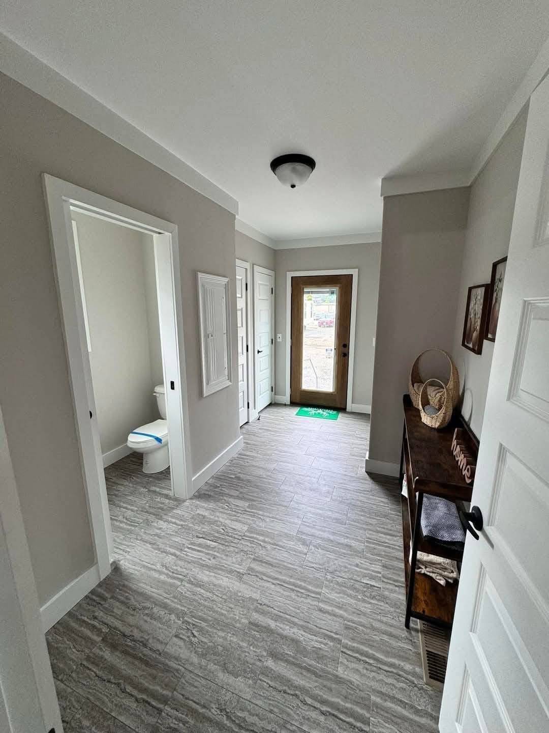 Narrow hallway with gray tiled flooring, a wooden entrance door at the end, a side table with wicker baskets on the right, and an open bathroom door on the left.
