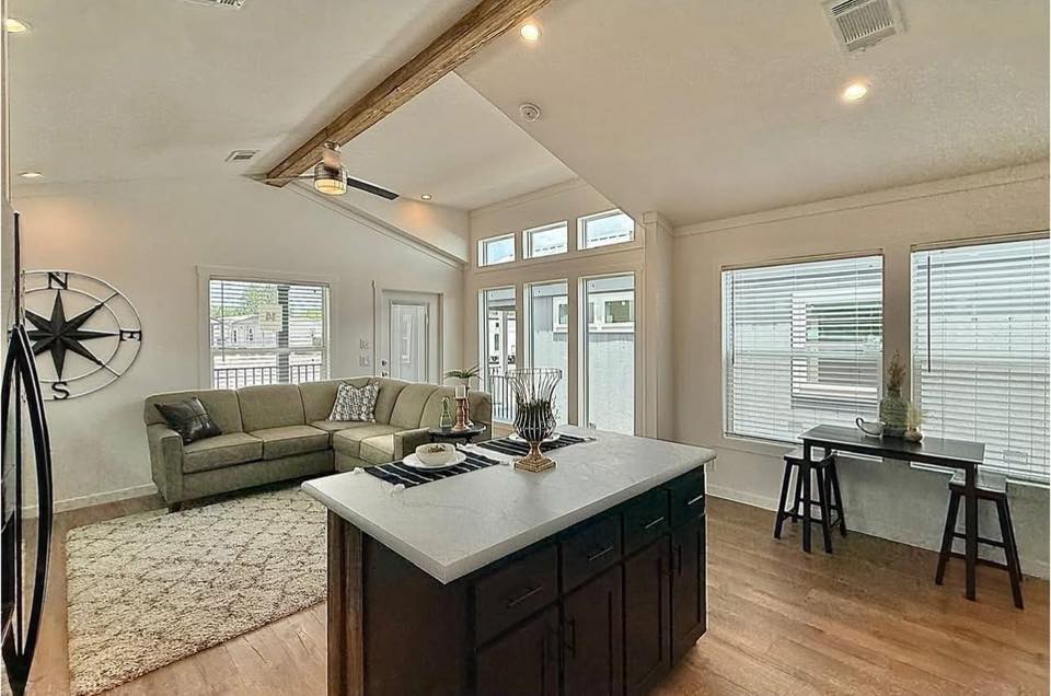 Spacious modern living room with high ceilings, light wood floors, and a wooden beam. Features a green sectional sofa, large island, and windows for natural light. A decorative compass adorns the wall.