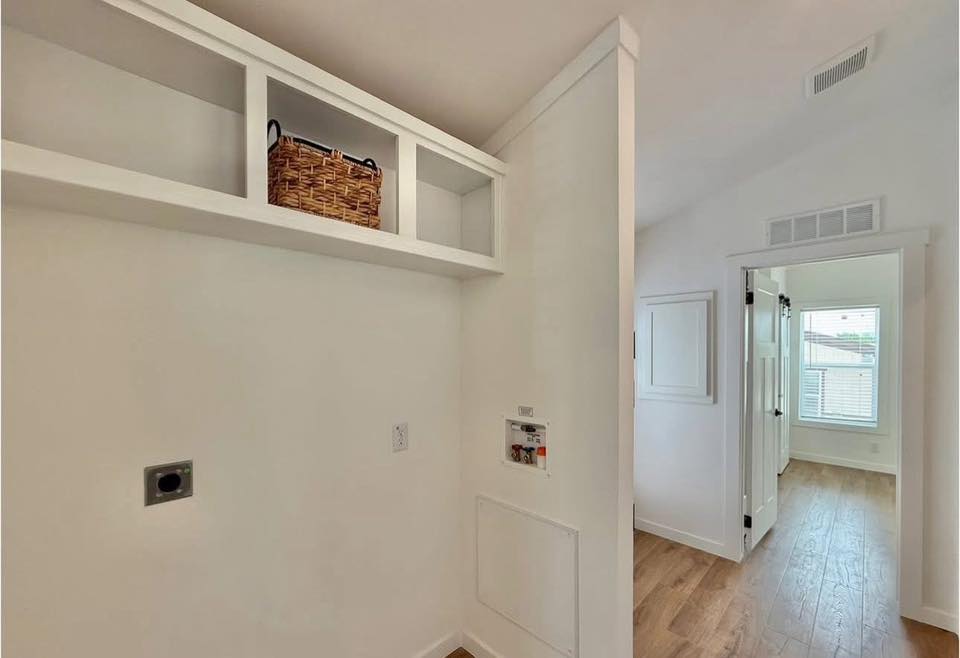 Laundry room with white walls, open shelves holding a wicker basket, wooden floor, and a view into an adjacent bright room through a doorway.