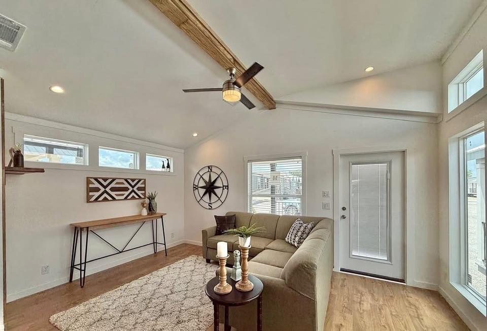 Bright living room with a modern beige sectional sofa, rug, and candles on a table. Decor includes a large wall clock and wood beam ceiling. Airy and inviting.