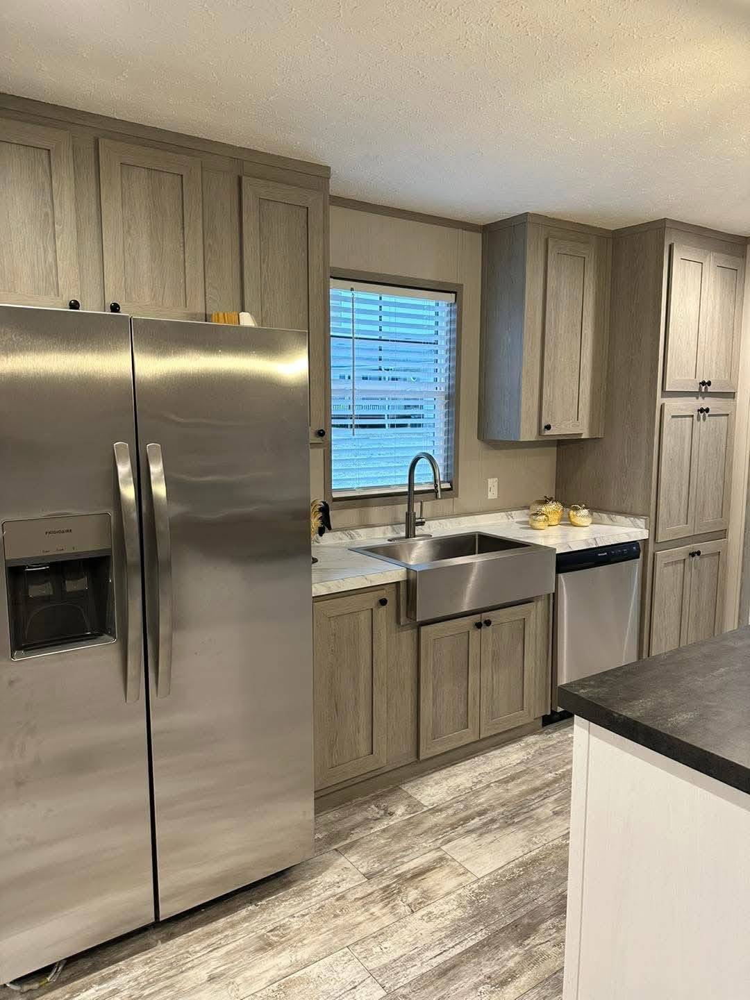 A modern kitchen with light wood cabinets, a stainless steel fridge, and an apron sink. A dishwasher is beside the sink under a window with blinds.