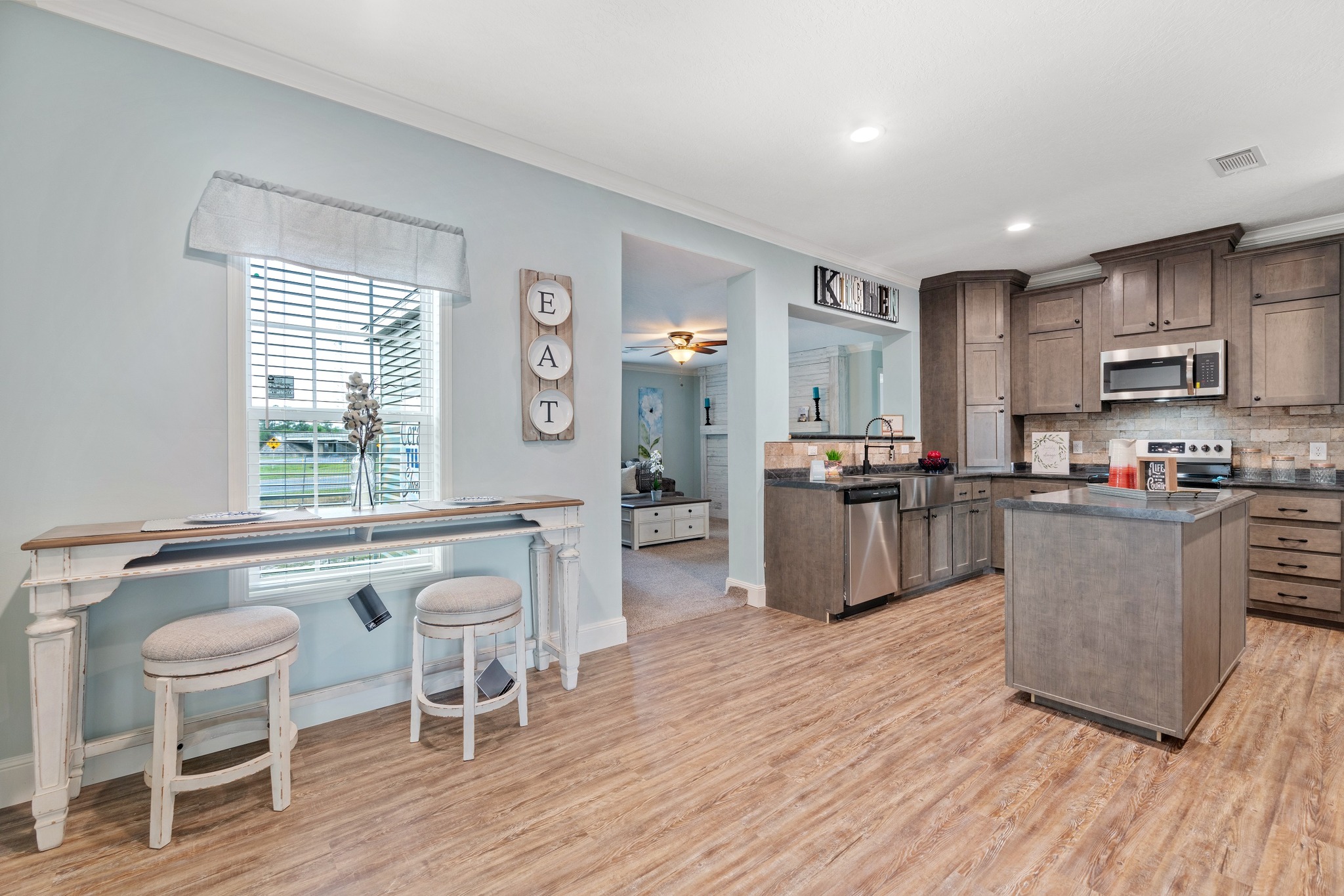 A modern kitchen with light wood flooring, gray cabinetry, and stainless steel appliances. A small dining nook with two stools is on the left. Cozy and inviting.