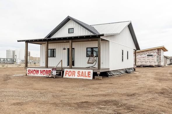 A small, white, single-story house with a porch and swing is displayed on a dirt lot. A large "Show Home For Sale" sign is in front. Overcast sky.