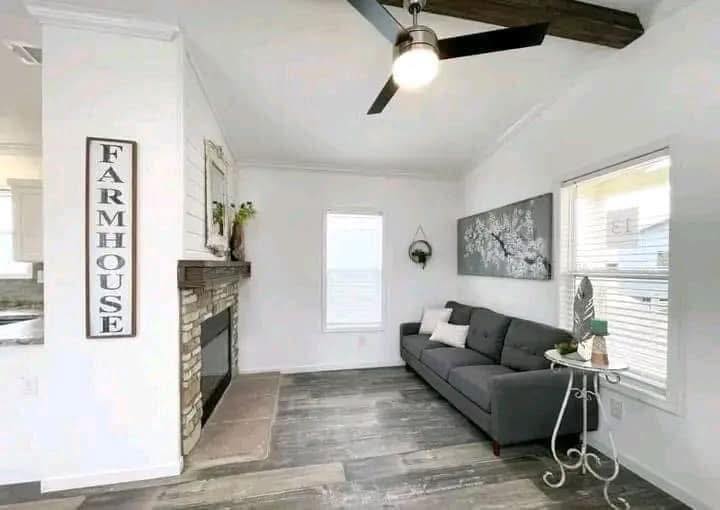 Bright living room with farmhouse decor. Features a dark gray sofa, rustic stone fireplace, leafy artwork, and wood accents, creating a cozy and airy ambiance.