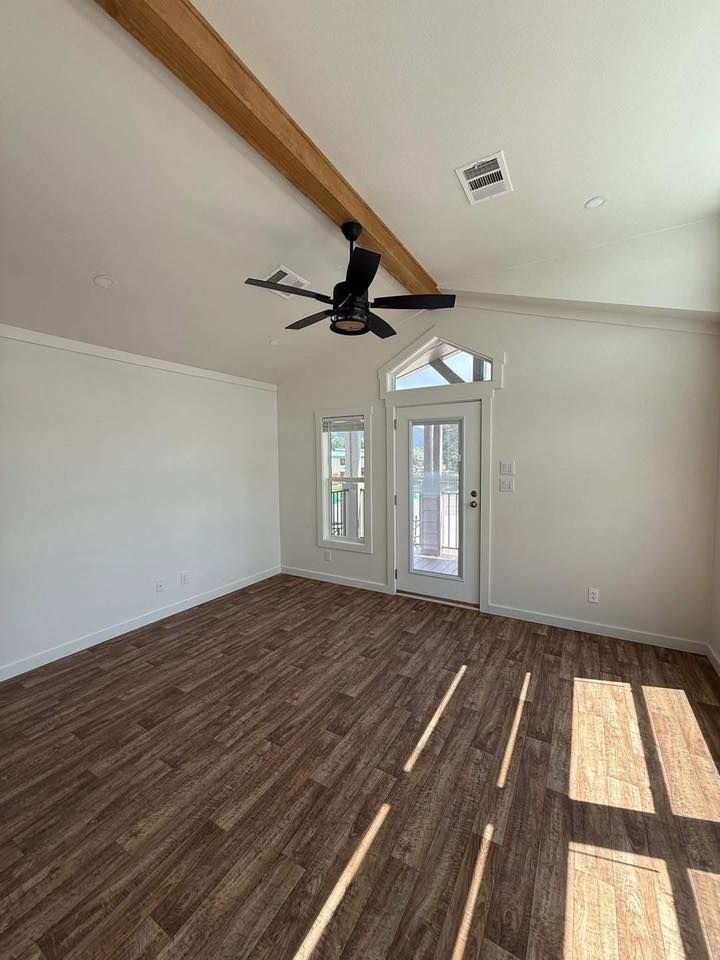 A bright, empty room with wood flooring and a vaulted ceiling. A single wood beam, ceiling fan, and sunlight through a glass door create a warm, inviting atmosphere.