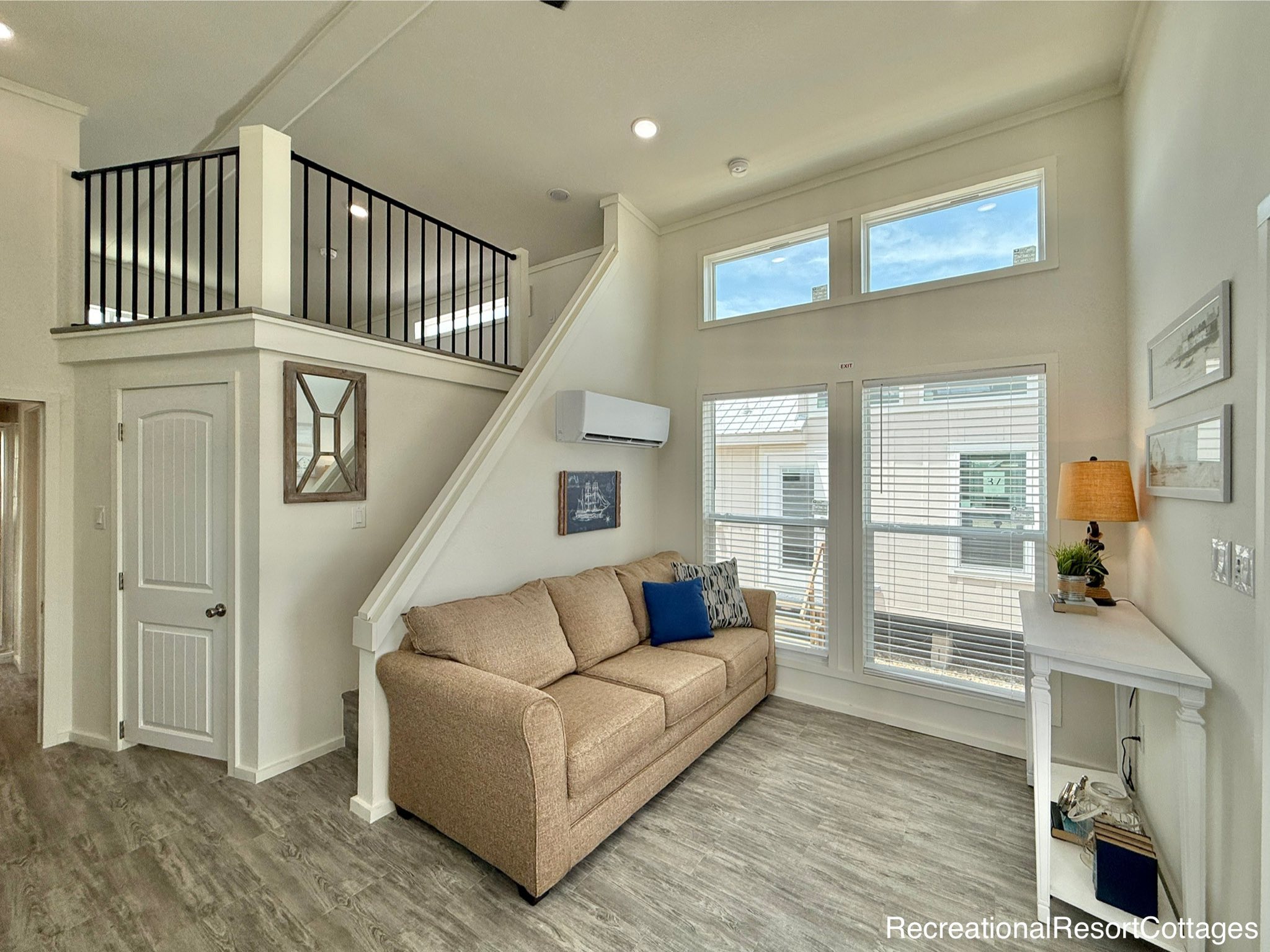 A cozy, modern living room with a beige sofa, decorative cushions, and a wall-mounted air conditioner. Large windows and a loft area add spaciousness.