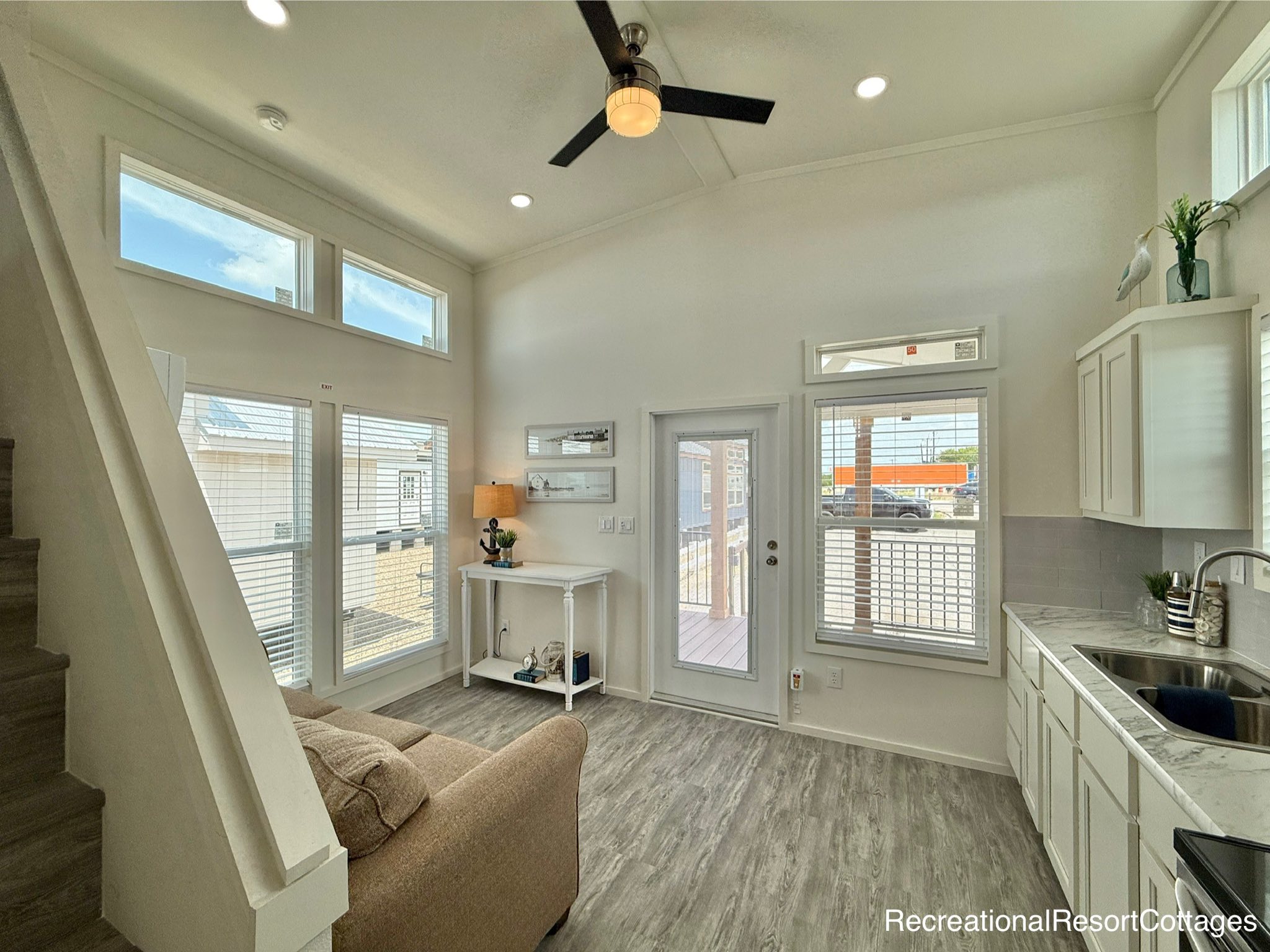 Bright living area with high ceilings, large windows, and modern decor. Features a beige couch, white cabinetry, gray flooring, and a ceiling fan, creating a spacious and airy atmosphere.